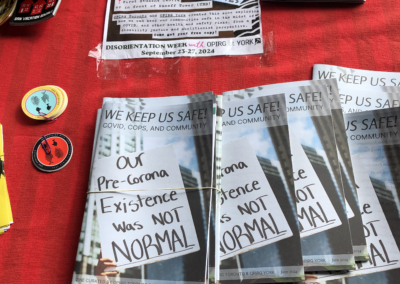 Red table cloth with several zines splayed out. The cover of the Zine says "We Keep Us Safe: Covid, Cops, and Community." The image on the cover shows a highrise in the background and a person's hand holding a hand-written sign that reads "Our Pre-Corona Existence was NOT normal."